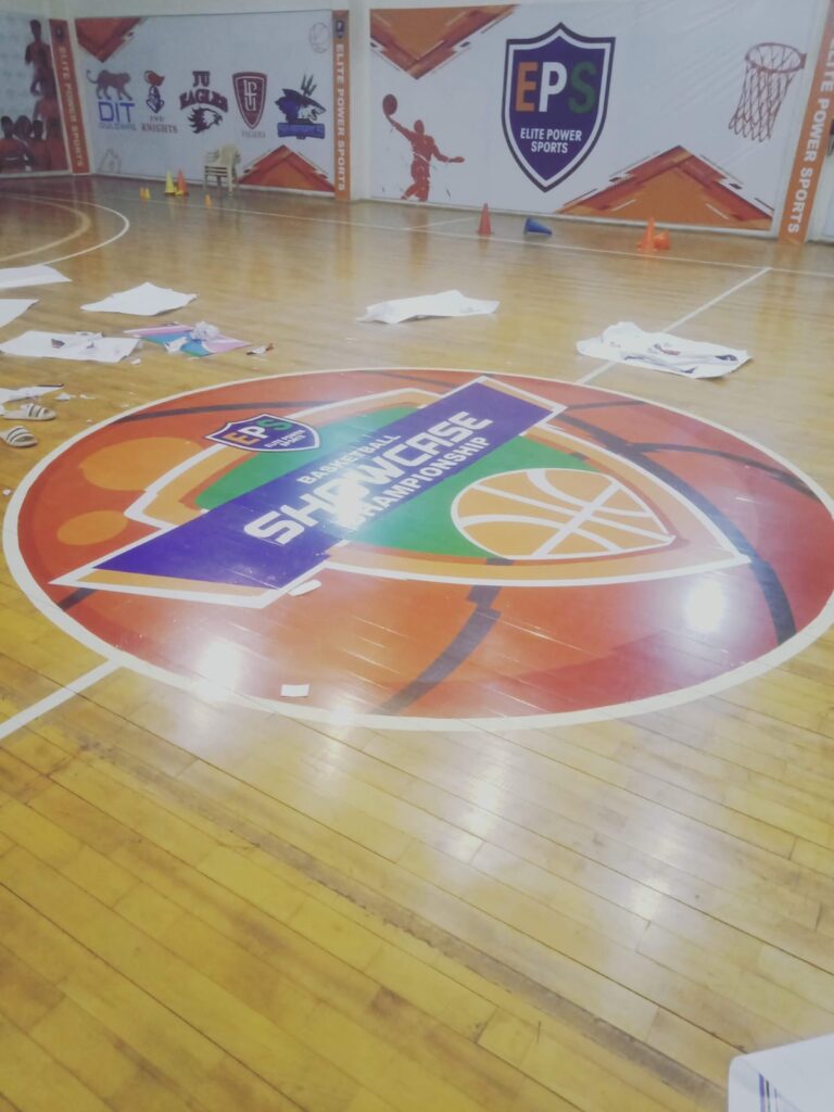 Basketball court with a vibrant Elite Power Sports Basketball Showcase Championship logo printed on the floor, surrounded by event branding and team logos.